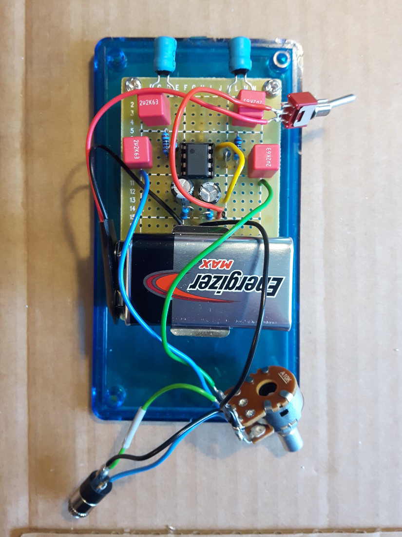 Photo of an electronic device without case; components on a circuit board: battery, resistors, capacitors (red), cables in different colors, switch. Placed on a cardboard.